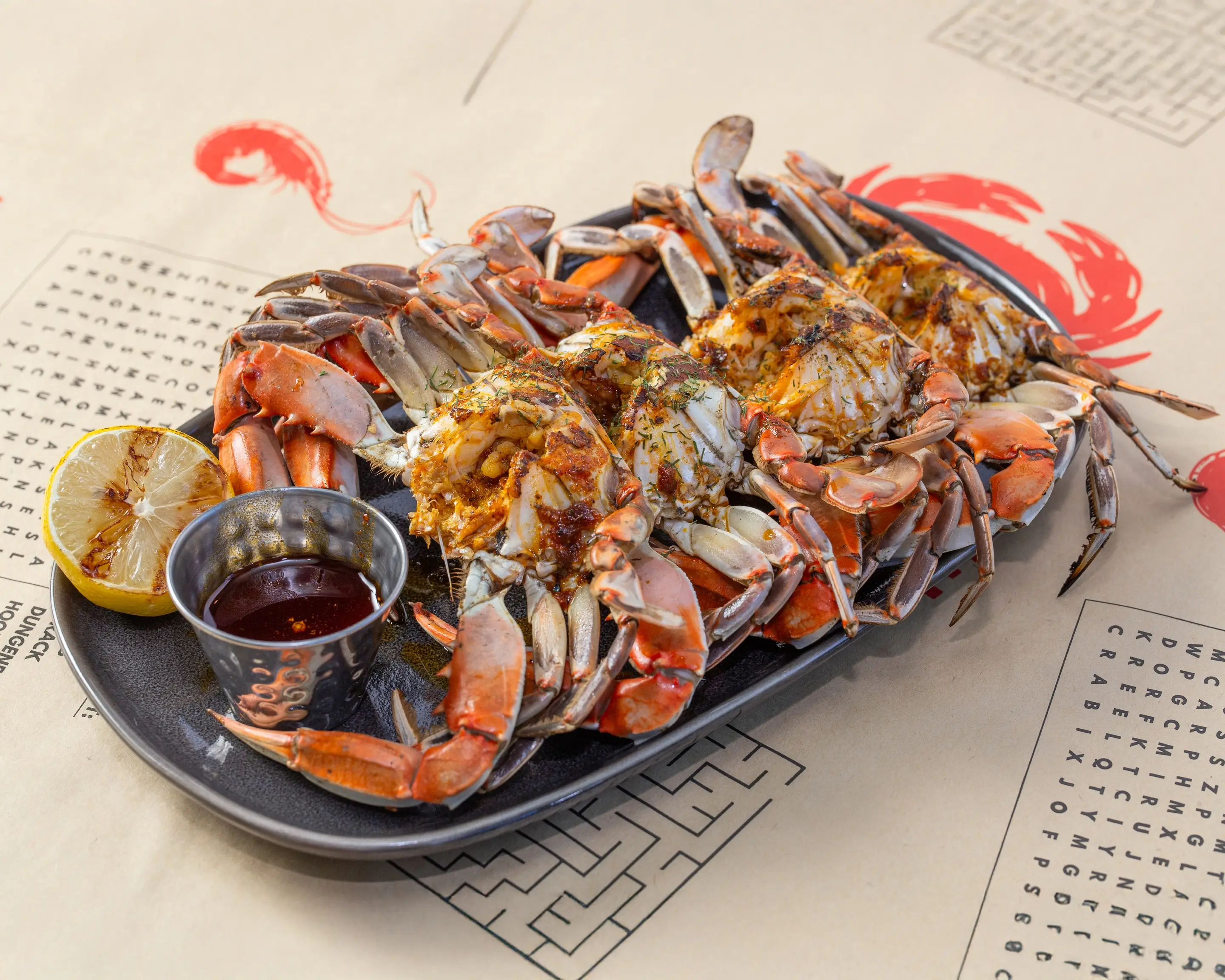 Halved blue crabs on a black platter, shells brushed with butter and flecked with herbs and Cajun seasoning, served with a charred lemon half and a tin of dipping sauce.