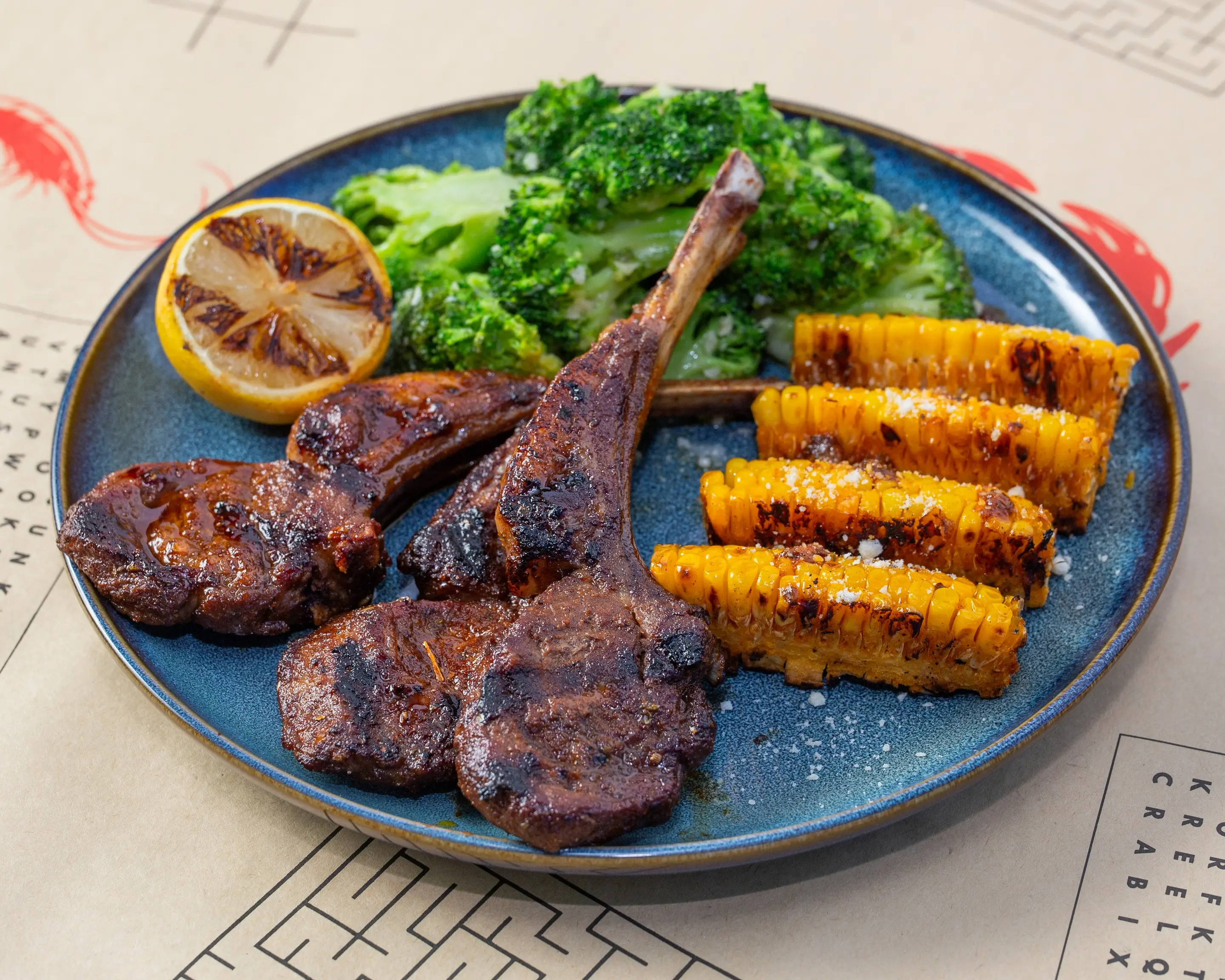 Grilled lamb chops on a blue stoneware plate, charred at the edges and served with garlic broccoli, charred corn ribs and a blackened lemon half.