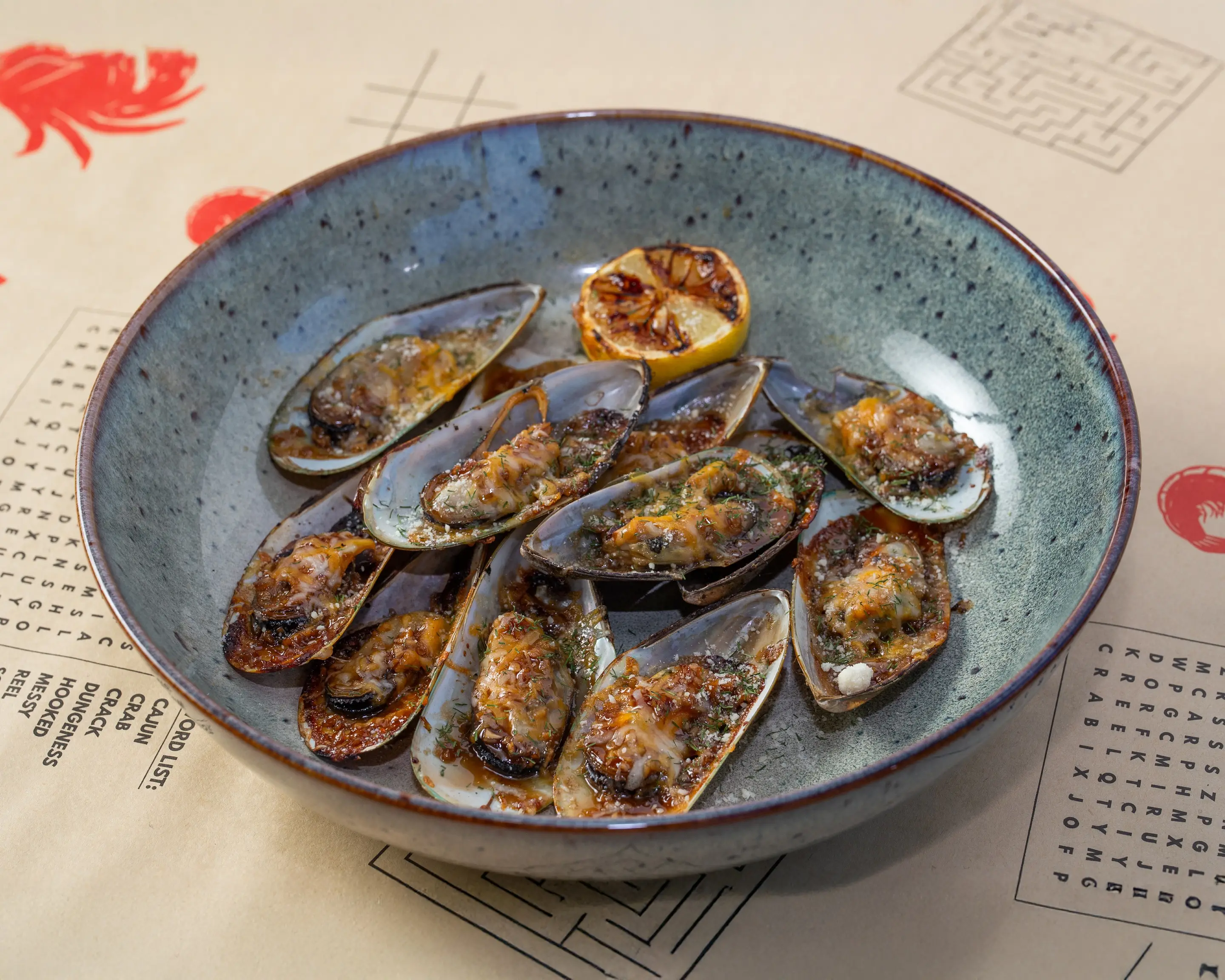 Mussels on the half shell filling a speckled stone bowl, grilled in garlic butter with herbs until the sauce catches, with a charred lemon half tucked at the back.