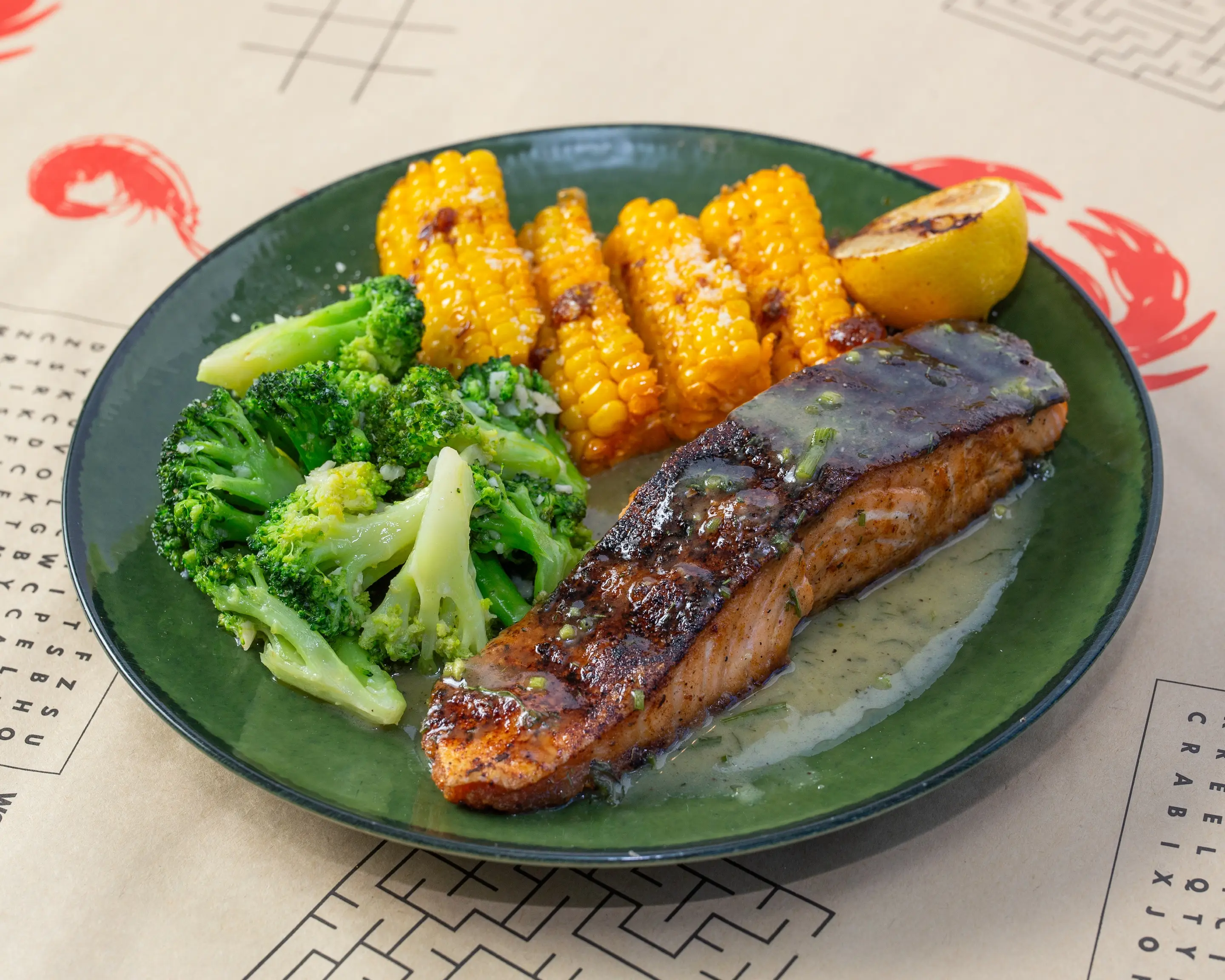 A salmon fillet grilled dark at the edges and glazed, resting in a butter sauce on a deep green plate with garlic broccoli, charred corn ribs and a wedge of lemon.