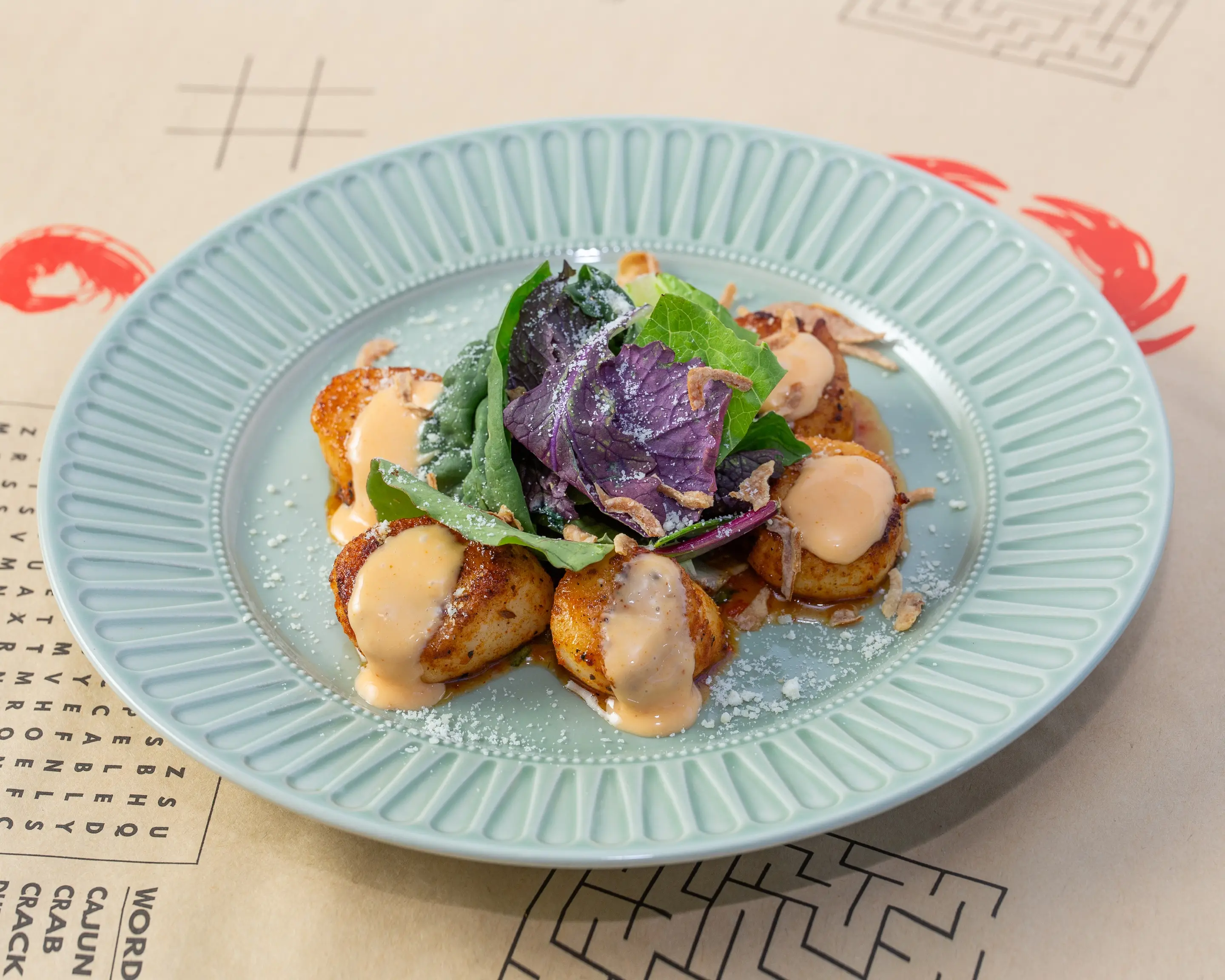 Six seared scallops arranged on a pale ribbed plate under a creamy pepper sauce, topped with dressed salad leaves and crisp fried shallots.